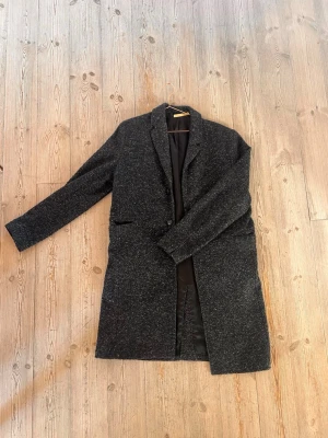 Ullrock Whyred  - Stilren mörkgrå kappa i ullmix med klassisk krage och två fickor framtill. Kappan har rak passform och helsvart foder, perfekt för kyligare dagar. Enkelknäppt design ger en tidlös look.