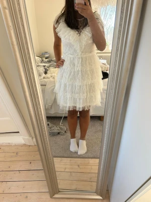 Vit spetsklänning med volanger - Söt vit kortklänning i spets med v-ringning och volangdetaljer. Klänningen har korta ärmar och flera lager spets som ger en romantisk känsla. Perfekt för sommarens alla tillfällen och har en feminin siluett.