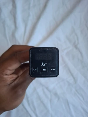 Svart FM-sändare till bil från KS - En svart FM-sändare till bil från KS med digital display och knappar för kanalbyte och uppspelning. Sändaren kopplas in i bilens cigarettuttag och har en kompakt, rektangulär form i plast. Perfekt för att spela musik via bilens radio.