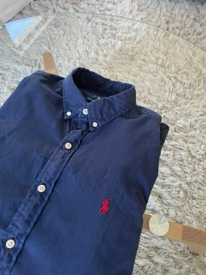 Polo Ralph Lauren Navy - Klassisk Skjorta från Ralph Lauren I mycket fint skick.  Skick: Utmärkt, inga skador eller skavanker såsom hål eller fläckar.  Mått: • Axel - Axel: 44cm • Axel - Midja: 61cm • Armhåla - Armhåla: 46cm • Axel - Ärmslut: 63cm  Detaljer • Tillverkad i 100% Bomull • Ikonisk Marinblå färg • Broderad logga på vänster sida av bröstet • Luktfri  Nypris: 1995kr - Mitt Pris: 449kr Skickas inom 24 timmar efter köp! 