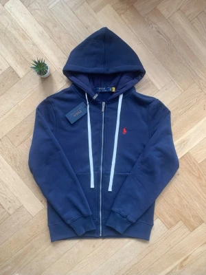 Mörkblå hoodie från Polo Ralph Lauren - Snygg mörkblå hoodie med dragkedja från Polo Ralph Lauren. Den har vit dragsko, huva och klassisk röd logga broderad på bröstet. Mjukt material och ribbade muddar vid ärmslut och nederkant. Perfekt för en chill look.