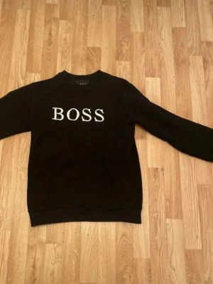 Svart sweatshirt från BOSS - Svart sweatshirt från BOSS med vit broderad logga på bröstet. Klassisk rund halsringning och långa ärmar. Perfekt för en avslappnad och stilren look.