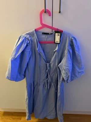 Ljusblå kortklänning från 157 - Supersöt ljusblå kortklänning från 157 med puffärmar och snörning framtill. Klänningen har en avslappnad passform och är gjord i ett mjukt bomullsmaterial. Perfekt för sommarens alla tillfällen!