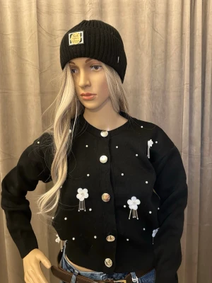 Svart kofta med pärldetaljer och blommor S-M - Trendig svart kofta med stora silvriga knappar, små pärldetaljer och vita blommor med hängande tofsar framtill. Rund halsringning och ribbade muddar. Perfekt att styla med jeans för en cool look.