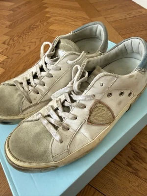Vita Philippe Model sneakers i läder i strl 37 - Säljer ett par vita Philippe Model sneakers i läder med grå detaljer och klassisk låg silhuett. Skorna har vita snören, gummisula och den ikoniska sköldloggan på sidan. Perfekta för dig som gillar stilrena och tidlösa sneakers. Det ena parets snören är lite trasiga och borde  bytas ut! Därav det låga priset! Nypris ca 4000
