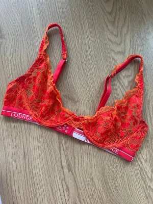 Lounge BH - Säljer en snygg röd/orange luxe spets-bh från Lounge med blommigt mönster och justerbara axelband. Bh:n har en elastisk resår med logga och är tillverkad i spets som ger en feminin look. Storlek 65C ❤️