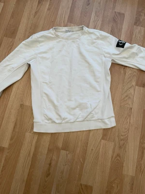 Vit sweatshirt från Calvin Klein - Snygg vit sweatshirt från Calvin Klein med rund halsringning och ribbade muddar. Tröjan har en svart patch med CK-logga på ena ärmen för en clean och modern look. Perfekt till jeans eller joggers för en avslappnad stil.