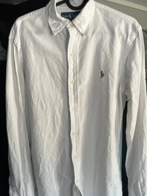 Vit skjorta från Ralph Lauren - Klassisk vit skjorta från Ralph Lauren med broderad logga på bröstet. Skjortan har lång ärm, knappar framtill och krage. Storleken är S, pris endast 499kr! Hör gärna av er vid frågor och funderingar!