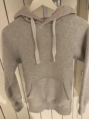 Ljusgrå hoodie med huva och ficka - En stilren ljusgrå hoodie med huva och dragsnören. Hoodien har en klassisk känguruficka framtill och ribbade muddar vid ärmar och nederkant. Tillverkad i mjukt sweatshirtmaterial, perfekt för en avslappnad look.
