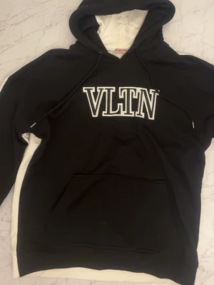 Svart VLTN hoodie från Valentino - Snygg svart hoodie från Valentino med stor vit VLTN-logga på bröstet. Hoodien har huva med dragsko, magficka och vita detaljer på insidan av huvan. Perfekt för dig som gillar streetwear och vill ha något stilrent men ändå trendigt.