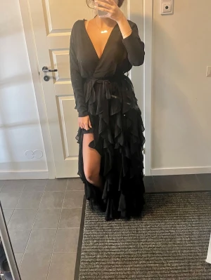 Svart långklänning med volanger - Säljer en elegant svart långklänning med djup v-ringning och volanger som ger en dramatisk look. Perfekt för festliga tillfällen! Klänningen har långa ärmar och en lös passform som gör den bekväm att bära hela kvällen. Även en lång slits vid benen. Passar även 36.