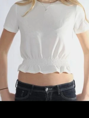 Vit croppad topp med resår - Säljer en vit croppad topp med korta ärmar och resår i midjan som ger en snygg rynkad effekt. Toppen har en enkel och fräsch look med lätt volangkant nedtill. Perfekt till jeans eller kjol för en trendig stil.
