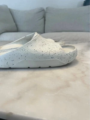 Vita Jordan sandaler med stänk - Säljer ett par vita sandaler från Jordan med svart stänkmönster. Tillverkade i ett mjukt och bekvämt syntetmaterial med präglad Jumpman-logga på fotbädden. Slip-on modell med modern, chunky sula och sportig känsla.