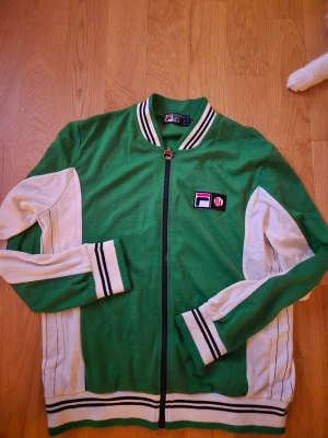 Grön och vit ziptröja från FILA BJ - Säljer en retroinspirerad ziptröja från Fila i grönt och vitt med svarta och vita ränder på muddar och krage. Tröjan har dragkedja framtill och broderad Fila-logga på bröstet. Perfekt för en sportig och avslappnad stil. Storleken är XL och det finns tecken på användning men i sparsamt skick. Finns i Hässelby. 