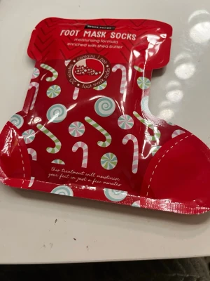 Foot Mask Socks med sheasmör - Roliga fotmask-strumpor från Sence i röd förpackning med godis- och polkagrisdesign. Innehåller återfuktande formula berikad med sheasmör som ger mjuka och återfuktade fötter. Perfekt för en snabb och enkel fotvårdsrutin hemma.