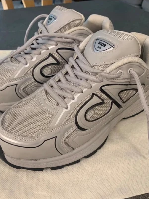 Grå Dior B30 sneakers - Säljer ett par grå Dior B30 sneakers med coola svarta detaljer och meshpaneler för extra andningsförmåga. Skorna har en chunky sula och tydlig Dior-logga på sidan. Perfekta för dig som gillar streetwear och exklusiva märken.