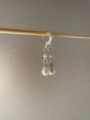 Silvriga örhängen med vita pärlor - Säljer ett par stilrena örhängen i silverfärg med droppformade vita pärlor och små runda pärldetaljer. Örhängena har en enkel och elegant design som passar perfekt till många olika looks. Materialet ser ut att vara metall och pärlor. Egengjorda 