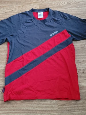 Adidas röd och grå t-shirt - Säljer en cool t-shirt från Adidas med diagonal design i rött och mörkgrått. T-shirten har korta ärmar och loggan på bröstet. Materialet är mjuk bomull och passformen är normal. Perfekt för dig som gillar sportiga och stilrena plagg.