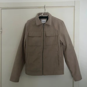 Beige mockajacka från Jack & Jones - Snygg beige mockajacka från Jack & Jones med raka linjer, klassisk krage och två stora bröstfickor med lock. Jackan har dragkedja framtill och långa ärmar. Perfekt för dig som gillar stilren och enkel design. Xs passar även S