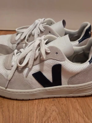 Vita Veja sneakers med blå detaljer - Snygga vita Veja sneakers med marinblå V-logga på sidan och blå detalj vid hälen. Ovandelen är i mesh och mocka, med vita skosnören och klassisk låg profil. Perfekta för en clean och stilren look.