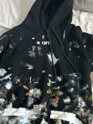 Svart hoodie från Off-White - Säljer en svart hoodie från Off-White med färgstänk och vit text på bröstet. Hoodien har justerbar huva med snören och vita detaljer på fickorna. Materialet är mjukt och bekvämt, perfekt för en streetwear-look.