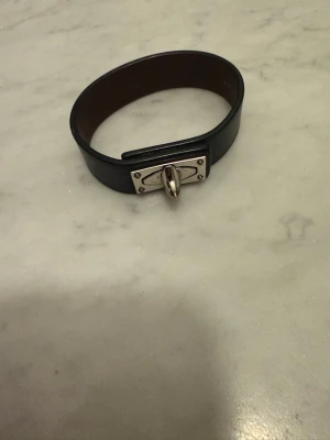 Svart läderarmband med metallspänne - Snyggt svart armband i läder med ett unikt silverfärgat metallspänne framtill. Armbandet har en stilren och modern design med bred rem och detaljer i metall som ger en exklusiv känsla. Perfekt accessoar för att lyfta din outfit. Strl M 