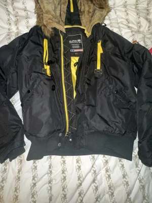 Svart bomberjacka Alpha Industries - Snygg svart bomberjacka från Alpha Industries med gul dragkedja och detaljer. Jackan har en stor huva med fuskpäls, ribbade muddar och flera fickor framtill. Insidan är gul och materialet är slitstark nylon. Perfekt för dig som vill ha en streetwear-look.