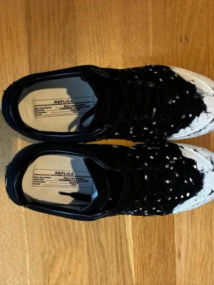 Masion margiela. svarta sneakers - Gats. sneakers i svart med vit färgstänk och slitna detaljer. Ovandelen är i tyg och mocka, med svart snörning och vit tå. Sulan är svart med rutmönster undertill. Unik och konstnärlig design som sticker ut.