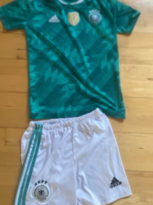 Tysklands fotbollsställ Adidas - Säljer ett Tysklands fotbollsställ från Adidas med grön mönstrad tröja och vita shorts. Tröjan har geometriskt mönster, vita detaljer och Tysklands landslagsmärke. Shortsen är vita med gröna ränder och Adidas-logga. Perfekt för fotbollsträning eller match.