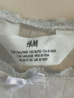 Vit långärmad topp från H&M - Söt vit långärmad topp från H&M med spetsdetaljer vid halsringningen och en liten rosett framtill. Tröjan har diskret ribbad struktur och är i mjukt material, perfekt för en enkel men gullig look.