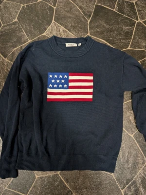 Mörkblå stickad tröja från RYVLS stl 158-164 - Mörkblå stickad tröja från RYVLS med amerikansk flagga broderad på bröstet. Tröjan har rund halsringning och ribbade muddar vid ärmslut och nederkant. Perfekt för dig som gillar klassisk och stilren design.