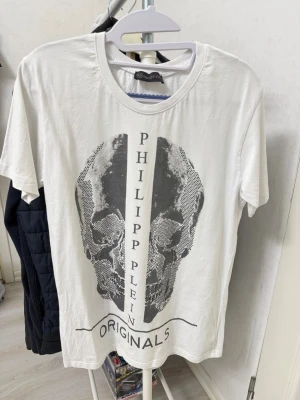 Vit Philipp Plein t-shirt med döskalle helt ny storlek S  - Säljer en vit t-shirt från Philipp Plein med ett stort svart döskalletryck framtill och texten 'ORIGINALS'. T-shirten har rund hals och korta ärmar. Perfekt för dig som gillar streetwear och vill sticka ut med en unik design. Nyskick i storlek s, aldrig använd. 