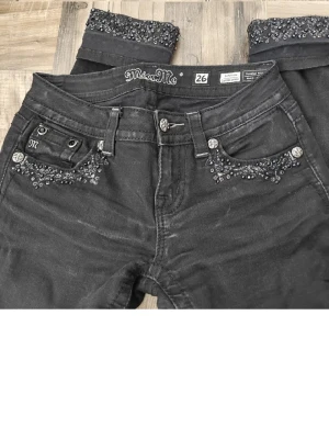 Svarta Miss Me jeans  - Snygga svarta jeans från Miss Me med dekorativa strassdetaljer på framfickorna och uppvikta benslut med extra bling. Klassisk femficksmodell och låg midja. Perfekta för dig som vill sticka ut med lite extra detaljer.