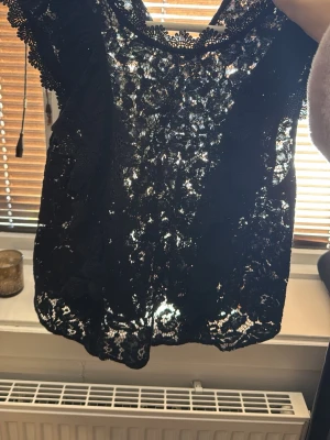 Svart spetsblus med volangdetaljer - Supersnygg svart blus i spets med volangdetaljer längs axlarna. Blusen är ärmlös och har ett transparent, blommigt spetsmönster som ger en cool och elegant vibe. Perfekt att styla med jeans eller kjol för en trendig look.