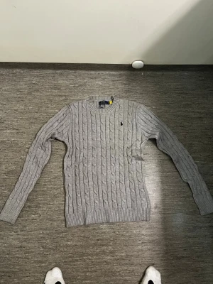 Ljusgrå kabelstickad tröja Polo Ralph Lauren - Snygg ljusgrå kabelstickad tröja från Polo Ralph Lauren. Tröjan har rund halsringning, ribbade muddar och den klassiska lilla logotypen broderad på bröstet. Tillverkad i mjukt material som passar perfekt till lager på lager.