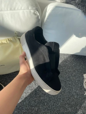 Svarta sneakers från Axel Arigato - Stilrena svarta sneakers från Axel Arigato i mocka med vit sula. Klassisk låg modell utan snörning, med diskret guldfärgad logga på sidan. Perfekta för dig som gillar minimalistisk och clean stil.