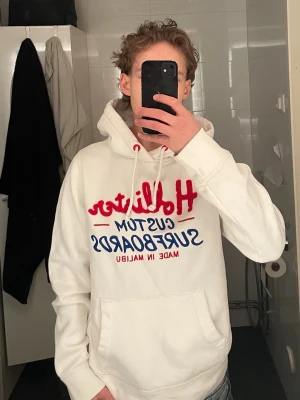 Vit Hollister hoodie med tryck - Snygg vit hoodie från Hollister med rött och blått tryck på bröstet där det står 'Hollister Custom Surfboards Made in Malibu'. Klassisk känguruficka och dragsko i huvan. Perfekt för en avslappnad och trendig stil.