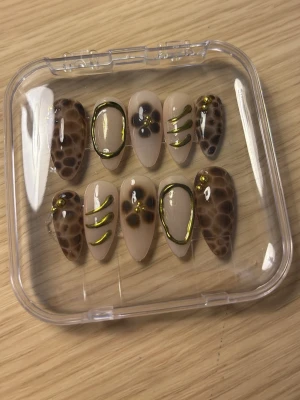 Bruna leopardmönstrade press-on naglar - Snygga press-on naglar i mandelform med beige bas, leopardmönster i brunt och detaljer i guld. Några naglar har guldfärgade linjer och små blommor för extra effekt. Perfekt för dig som vill ha en unik och trendig look. 10st naglar!!!