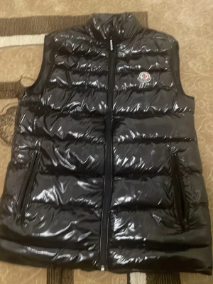 Moncler väst - Säljer en svart, glansig dunväst från Moncler med hög krage och dragkedja framtill. Västen har två fickor med dragkedja och Moncler-logga på bröstet. Perfekt för lager-på-lager och riktigt snygg till streetwear.