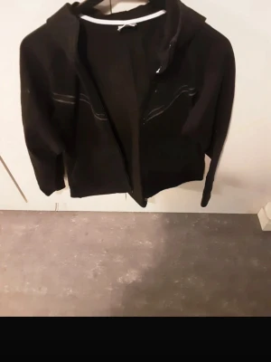 Svart hoodie med dragkedja - Svart hoodie med dragkedja framtill och huva. Klassisk modell med långa ärmar och två fickor på sidorna. Perfekt för en avslappnad och stilren look. Passar till det mesta och är enkel att matcha med andra plagg.