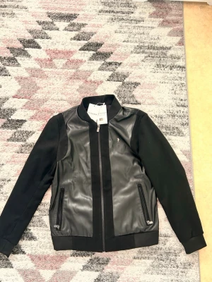 Svart skinnjacka från U.S. Polo Assn. - Snygg och helt oanvänd svart skinnjacka från U.S. Polo Assn. med svarta tygärmar och ribbade muddar. Jackan har två dragkedjeförsedda fick sopå framsidan och en liten silverfärgad logga på bröstet. Perfekt för dig som vill ha en stilren och modern look.