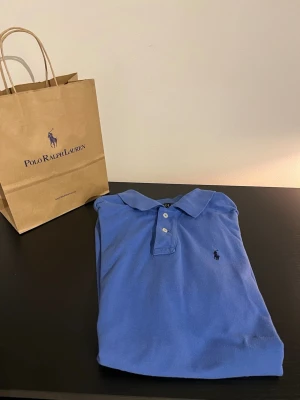 Blå pikétröja från Polo Ralph Lauren - Snygg blå pikétröja från Polo Ralph Lauren med klassisk krage och två knappar framtill. Tröjan har den ikoniska loggan broderad på bröstet och är tillverkad i mjuk bomull. Perfekt för en clean och stilren look. Skriv vid fler bilder eller om du har några frågor.