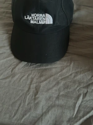 Svart keps The North Face Malmö - Snygg svart keps från The North Face med vit brodyr där det står 'Norra Läktaren Malmö'. Klassisk böjd skärm och justerbar passform. Perfekt accessoar för dig som vill ha en clean och stilren look med street-vibe.