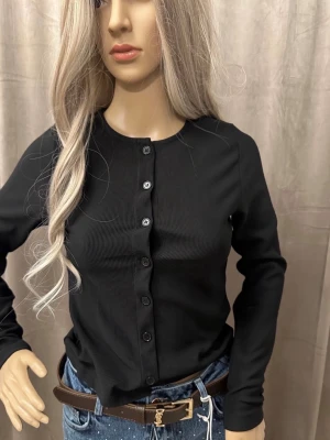 Svart ribbad kofta med knappar XS-S - Stilren svart ribbad kofta med rund halsringning och knäppning framtill. Långärmad modell i mjukt, stretchigt material som sitter snyggt på kroppen. Perfekt att matcha med jeans eller kjol för en trendig look.