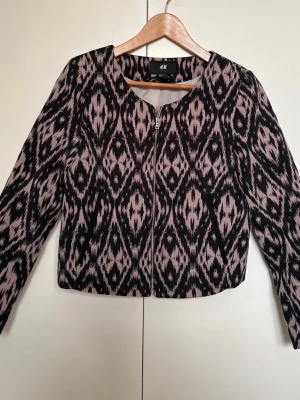 Mönstrad jacka från H&M - Säljer en svart och beige jacka från H&M i storlek 38 men sitter som en 34. Jackan har rundad halsringning, dragkedja framtill och är kort i modellen. Perfekt för att lyfta en enkel outfit med lite edge.