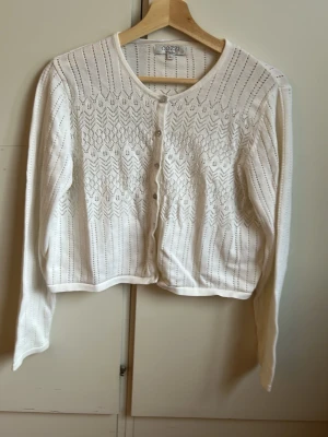 Vit stickad kofta  - Söt vit stickad kofta från Cozzi med genombrutet mönster och rund halsringning. Koftan har långa ärmar, knäppning framtill med små knappar och en kortare, rak passform. Perfekt att slänga över en topp för en fräsch look. 