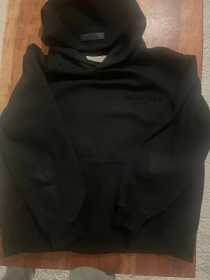 Svart Essentials hoodie med huva - Säljer en svart hoodie från Essentials med diskret logga på bröstet. Hoodien har en stor huva, ribbade muddar och en klassisk känguruficka. Perfekt för dig som gillar streetwear och enkel stil. Materialet känns mjukt och bekvämt.