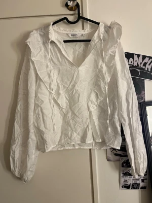 Vit volangblus från Kappahl - Snygg vit blus från Kappahl med volangdetaljer längs v-ringningen och axlarna. Blusen har lång ärm och är tillverkad i ett lätt, krinklat tyg som ger en avslappnad känsla. Perfekt att styla med jeans eller kjol för en trendig look.