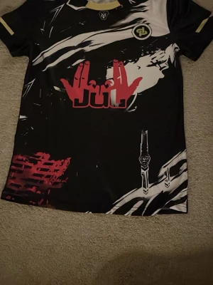 Svart grafisk t-shirt JUL logga - Unik svart t-shirt med grafiskt tryck i vitt, rött och gult samt JUL-logga och handmotiv på bröstet. T-shirten har korta ärmar och sportig känsla, perfekt för dig som gillar streetwear och statement-plagg. Materialet känns som polyester.