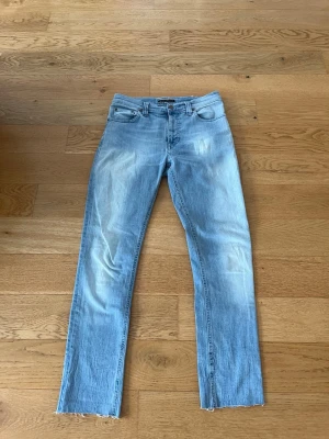 Ljusblå raka jeans från Nudie - Säljer ett par ljusblå raka jeans från Nudie med klassisk femficksmodell och råa, avklippta benslut. Jeansen har en avslappnad passform och är tillverkade i mjuk denim. Perfekta för en casual och trendig look.